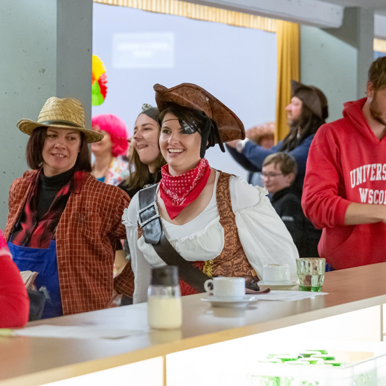 Eine Gruppe von Menschen in Kostümen steht um eine Theke. Eine Frau in einer Piratenhaube und einem Halstuch lächelt in die Kamera. Es gibt Tassen und Gewürze auf der Theke.