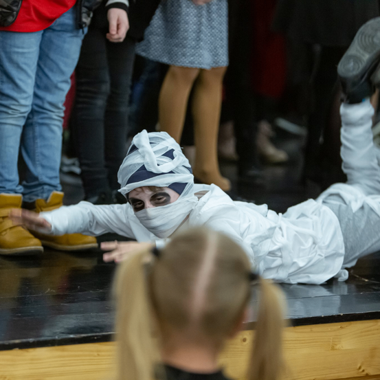 Eine Person in einem Mumienkostüm liegt auf dem Boden, umgeben von Zuschauern in einem schwach beleuchteten Raum.