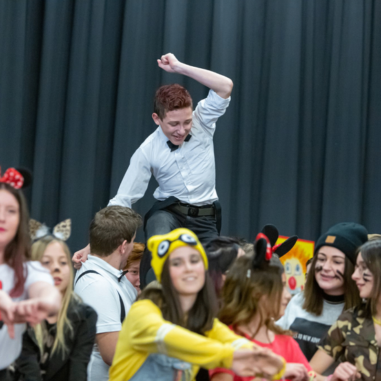 Ein Junge mit einer Fliege springt über einer Gruppe von Kindern, die teilweise Masken tragen, in der Luft. Eines der Mädchen in einer gelben Kostümierung lächelt im Vordergrund.