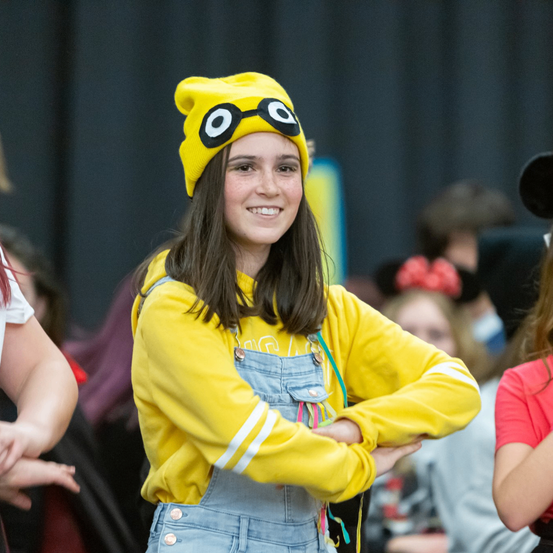 Ein lächelndes Mädchen in einem gelben Kostüm mit einem gelben Hut mit schwarzen Augen und einem Bogen auf dem Kopf, steht mit verschränkten Armen.