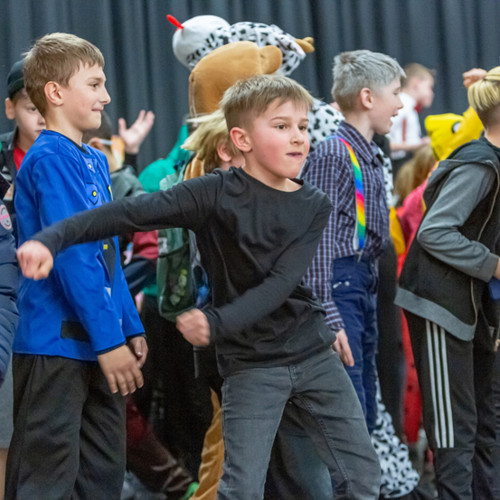 Eine Gruppe von Kindern bei einer Schulveranstaltung, einige tragen Kostüme, ein Junge in der Mitte schaut in die Kamera, während andere im Hintergrund verschwommen sind.