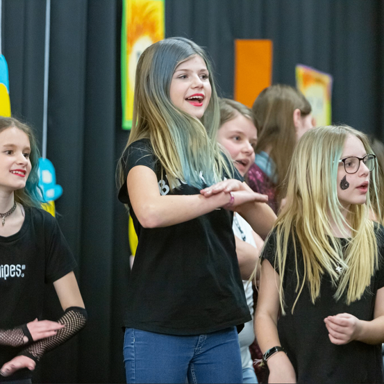 Eine Gruppe junger Mädchen mit verschieden gefärbtem Haar steht zusammen, möglicherweise bei einer Aufführung. Ein Mädchen hat ein Gesichtsdesign. Sie tragen schwarze T-Shirts.