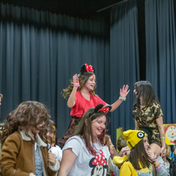 Mehrere Kinder in Kostümen, darunter Minnie Maus, tanzen fröhlich in einem Raum mit blauen Vorhängen. Ein Kind steht auf den Schultern eines anderen, und andere klatschen. Die Umgebung scheint ein Bühnen- oder Aufführungsbereich zu sein.