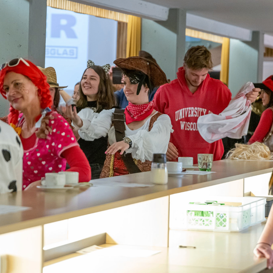 Mehrere Menschen in Kostümen versammeln sich an einer Theke, lächeln und halten Gegenstände. Eine Frau mit roter Perücke schaut einen Mann in einem roten University of Wisconsin Hoodie an. Dahinter ist eine Projektionsleinwand mit dem Wort 'Solar'.