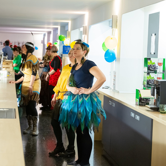 Eine Gruppe von Menschen in einem Flur, in Kostümen, stehen vor einem Tresen mit Luftballons und Kisten Wein.