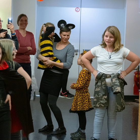 Eine Gruppe von Menschen ist in einem Raum, eine Person hält ein Kind, eine andere macht ein Foto, und ein Mädchen in einem Leopardendruckkleid. Hinter ihnen befindet sich ein Feuerlöscher und eine Tür.