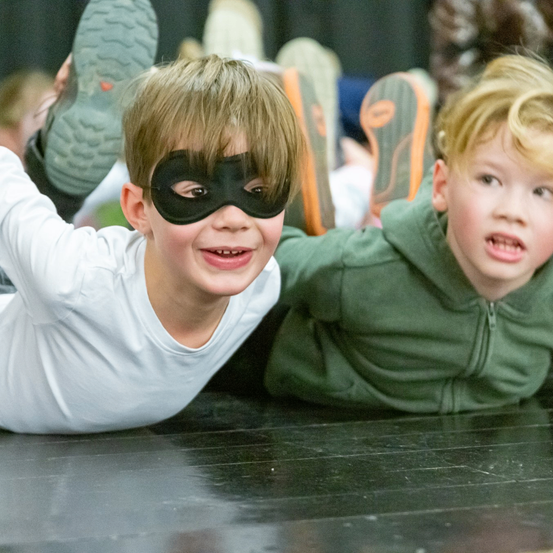 Zwei junge Jungen mit Masken, einer in Weiß und einer in Grün, liegen auf einem schwarzen Boden mit anderen dahinter.