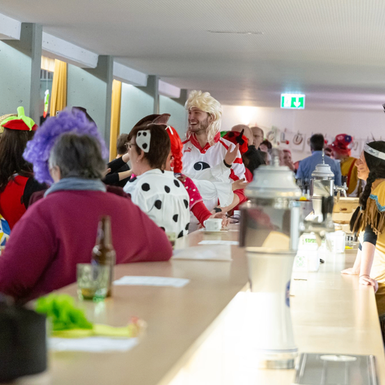 Eine Gruppe von Menschen in Kostümen sitzt an einer Bar. Einige tragen Hüte und andere haben Brillen. Eine Frau mit lila Haaren sitzt vorne.
