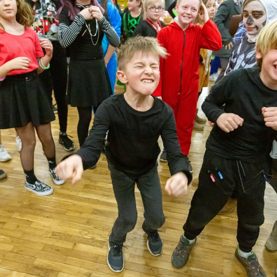 Mehrere Kinder tanzen und lachen auf einem Holzboden. Ein Junge vorne trägt schwarze Kleidung und Turnschuhe. Hinter ihm ein Junge in einem roten Kostüm, der lächelt. Ein anderer Junge in Schwarz lacht rechts.