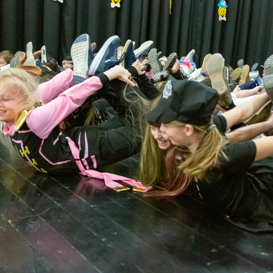 Zwei Mädchen sind auf einer Bühne, beide tragen schwarze Kleidung, eine mit einem rosa Langarmshirt. Sie spielen und lachen, liegen auf dem Boden mit vielen Schuhen um sie herum.
