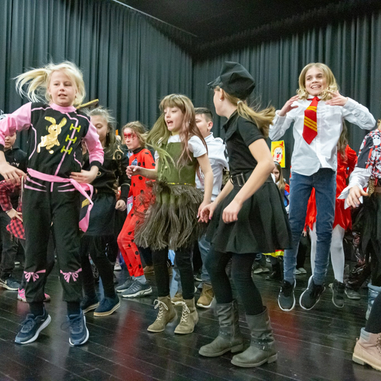 Mehrere Kinder in verschiedenen Kostümen führen einen Tanz auf einer Bühne mit schwarzem Hintergrund auf. Das Mädchen in Pink trägt ein Hasenkostüm und eine rosa Schleife.