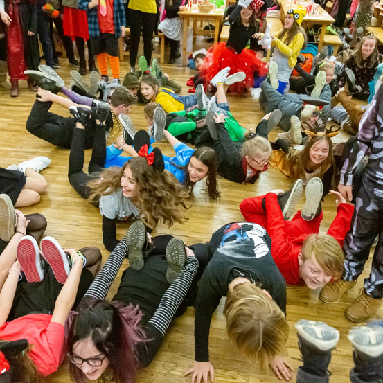 Eine Gruppe von Kindern in verschiedenen Kostümen, einige auf dem Boden liegend, scheinen an einer lustigen Aktivität teilzunehmen. Einige Erwachsene stehen hinter ihnen, und Tische mit Gegenständen sind in der Nähe.