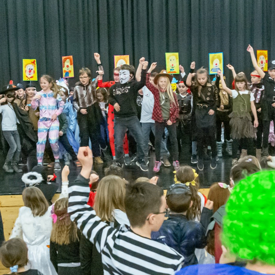 Eine Gruppe von Kindern in Kostümen tritt auf einer Bühne auf, einige halten bemalte Schilder hoch, und eine maskierte Person in der Mitte springt mit erhobenen Armen.