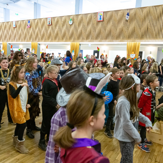Eine große Gruppe von Kindern in Kostümen ist in einem Saal versammelt. Einige tragen Hüte und Hasenohren, andere Halsketten und Brillen. Einige halten Luftballons.