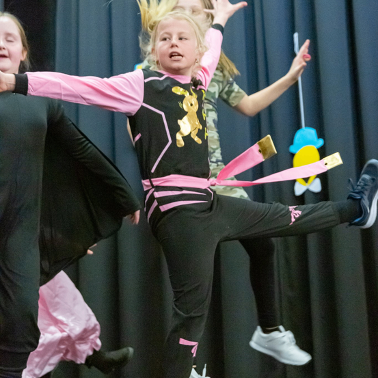 Ein Mädchen führt eine Gymnastikroutine auf der Bühne durch, trägt ein rosa-schwarzes Trikot mit einem goldenen Bärendesign. Eine andere Mädchen steht hinter ihr und beide haben ihre Hände erhoben.