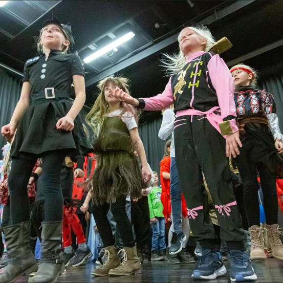 Kinder in verschiedenen Kostümen stehen auf der Bühne, eine Mädchen in einem schwarzen Kleid und Stiefeln schaut nach oben, ein Mädchen in einem grünen Kleid und Stiefeln schaut nach unten und ein Mädchen in einem pinken und schwarzen Kostüm hält eine Holzschwert.