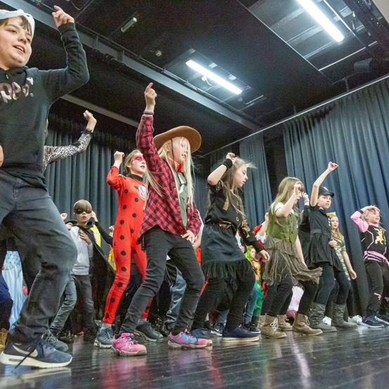 Eine Gruppe von Kindern in Kostümen führt eine Tanzroutine auf einer Bühne mit Lichtern und Vorhängen im Hintergrund auf.