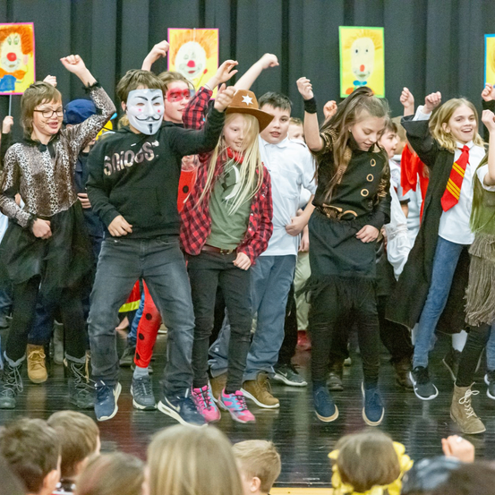 Eine Gruppe von Kindern in verschiedenen Kostümen und Masken tanzt auf einer Bühne mit farbenfrohen Hintergründen. Ein Kind trägt eine weiße Maske mit schwarzem Umriss.