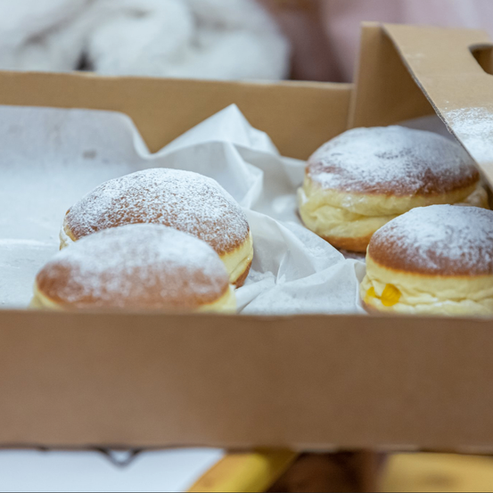 Eine Schachtel mit fünf puderzuckerbedeckten Donuts, auf weißem Papier, in einer Pappschachtel, mit verschwommenen weißen Objekten im Hintergrund.