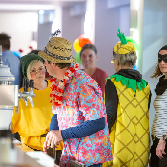 Eine Gruppe von Menschen in Kostümen und Hüten steht in einem Raum. Ein Mann trägt einen Ananas-Hut.