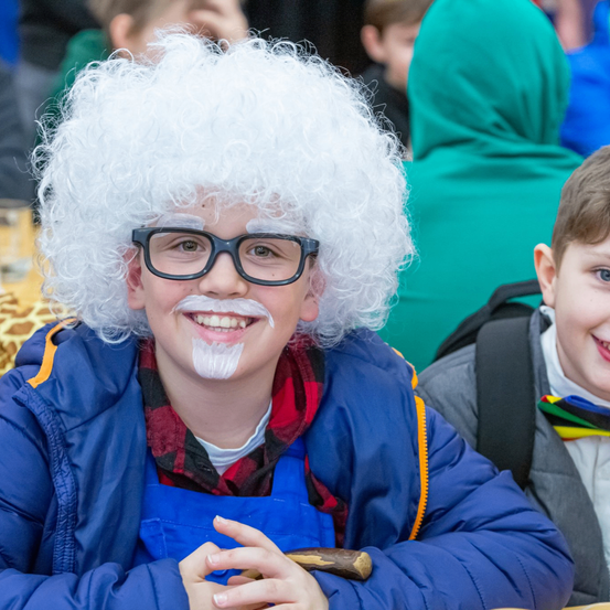 Ein junger Junge mit weißer Perücke und Brille hält einen Holzstab und lächelt. Ein anderer Junge mit einem Regenbogen-Schleife und einem Rucksack lächelt neben ihm.