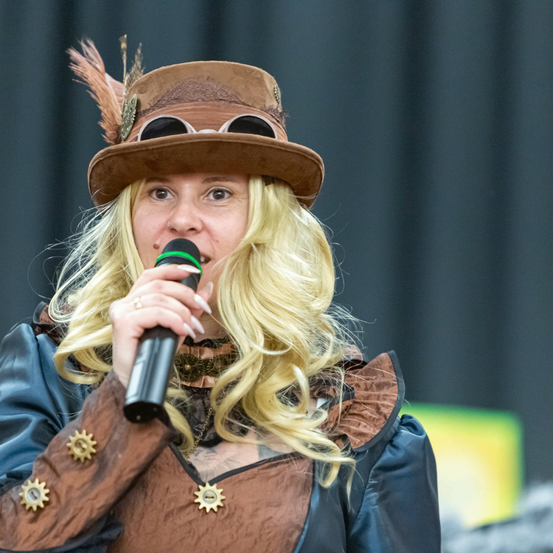 Eine Frau mit langen blonden Haaren, gekleidet in einem Steampunk-Kostüm, hält ein Mikrofon. Sie trägt einen braunen Hut mit Federn und eine Brille. Sie steht vor einer Bühne.