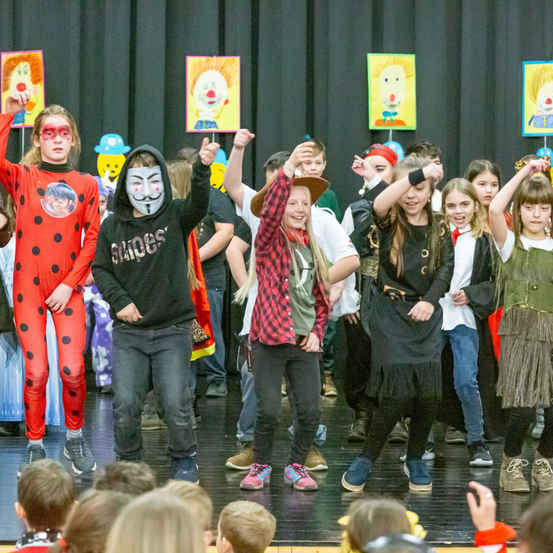 Eine Gruppe von Kindern tritt auf der Bühne in Kostümen auf. Ein Kind trägt ein rotes Kleid mit Punkten. Hinter ihnen hängen bunte Clown-Gemälde an der Wand.
