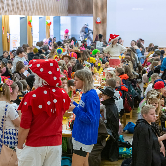 Ein Saal voller Menschen in Kostümen, die an Tischen sitzen, einige stehen, wobei eine Person in einem roten Pilzkostüm hervorsticht. Viele bunte Hüte und Dekorationen sind sichtbar, und die Szene ist lebhaft und festlich.