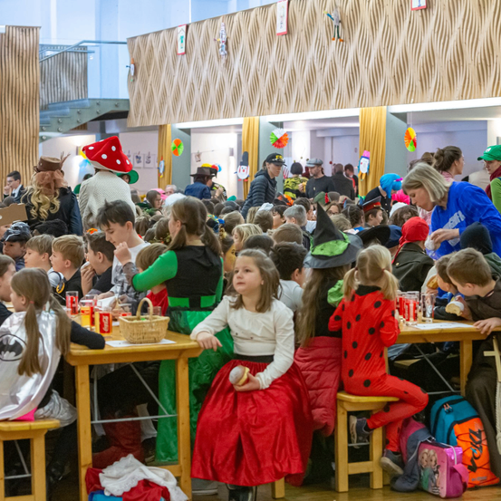Eine Menschenmenge von Kindern in Kostümen, darunter ein Mädchen in einem roten Kleid und ein anderes in einem Kostüm mit Punkten, sitzen um Tische herum bei einer Indoor-Veranstaltung. Sie sind von Erwachsenen umgeben, einige ebenfalls in Kostümen, und Dekorationen.
