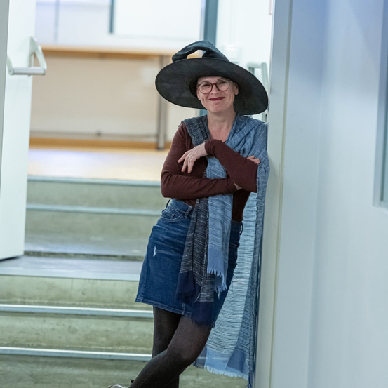 Eine Frau mit schwarzem Hut, Brille und Jeansrock lehnt sich in einem Flur mit Treppen an eine Tür.