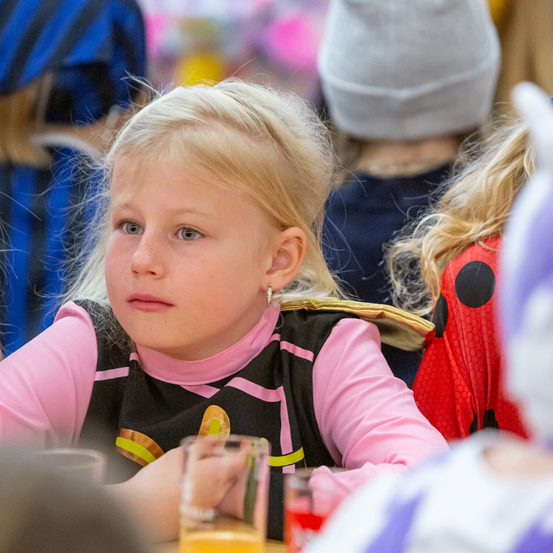 Ein junges Mädchen mit blonden Haaren, das ein rosa und schwarzes Kleid trägt, sitzt an einem Tisch mit einem Glas Orangensaft. Hinter ihr ist eine Person in einem blauen Shirt und eine Dame in einem roten Punktekleid zu sehen.