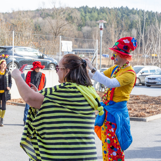 Ein Clown in leuchtenden Farben und einer Hut mit Blumen steht neben einer Frau in einem gestreiften Shirt und hält Scheren in einem Parkplatz. Dahinter sind Menschen in Kostümen und ein Auto zu sehen.