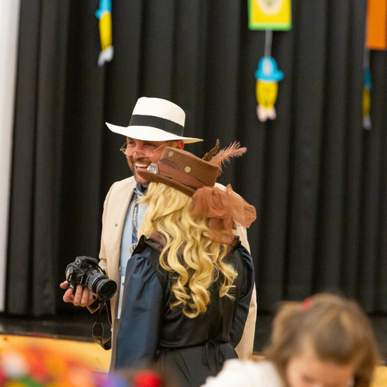 Ein Mann in einem weißen Hut fotografiert eine Frau mit einem braunen Hut und federigen Accessoires, während sie beide lächeln und vor einem schwarzen Vorhang mit bunten Dekorationen stehen.
