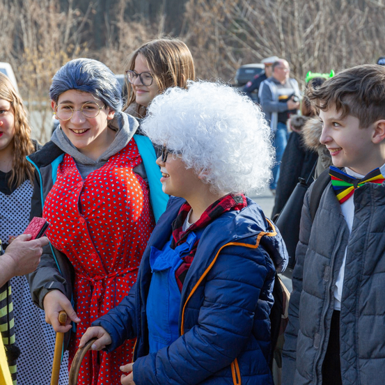Eine Gruppe von Menschen in Kostümen, eine Frau in einem roten Punktekleid, eine andere in einer weißen Perücke und ein Junge mit einem bunten Fliegenbinder, alle lächelnd und interagieren draußen.