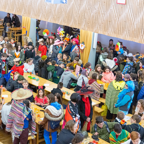 Eine große Gruppe von Menschen, wahrscheinlich Schüler, ist in einer Cafeteria versammelt. Sie sitzen an langen Holztischen, einige in Kostümen. Der Raum hat Holzbalken an der Decke und gelbe Vorhänge im Hintergrund. Viele Personen haben Getränke und Snacks vor sich.