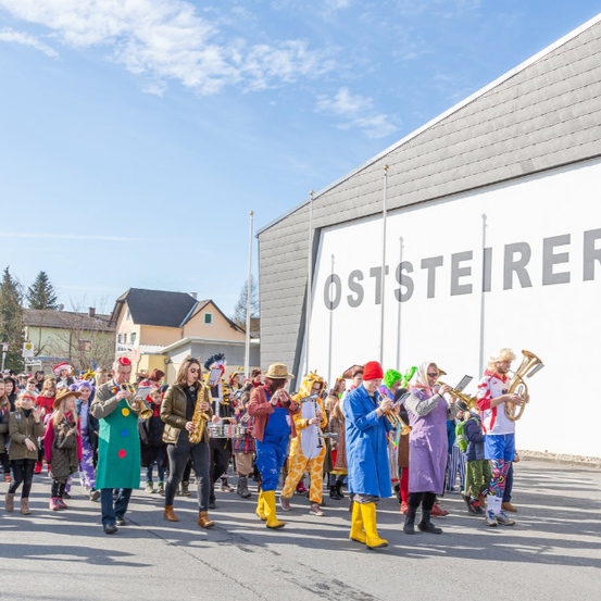 Eine Musikkapelle in bunten Kostümen spielt Instrumente vor einem Gebäude mit dem Wort Oststeierer darauf.