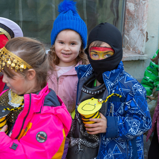 Drei Kinder in Kostümen lächeln vor einem Gebäude. Das Mädchen links trägt eine rosa Jacke und eine goldene Kopfbedeckung. Das mittlere Kind hat eine blaue Mütze und eine rosa Jacke. Der Junge rechts trägt eine schwarze Maske mit bemaltem Gesicht.