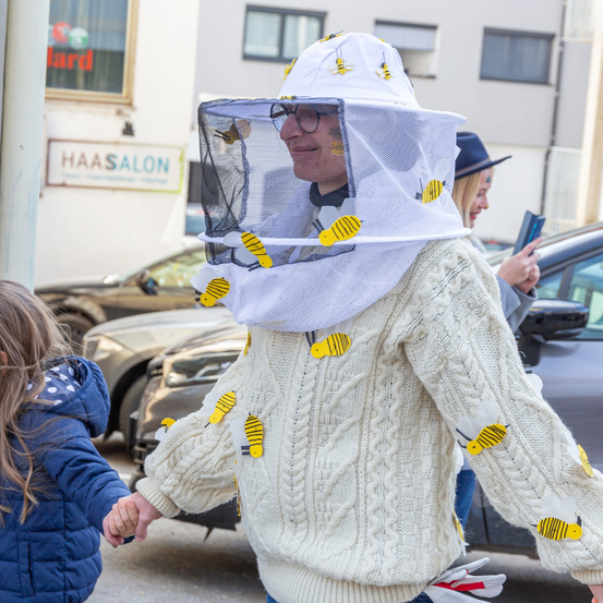 Ein Mann in einem von Bienen bedeckten Pullover und Hut geht mit einem Kind die Straße entlang, mit geparkten Autos und einem Gebäude im Hintergrund.