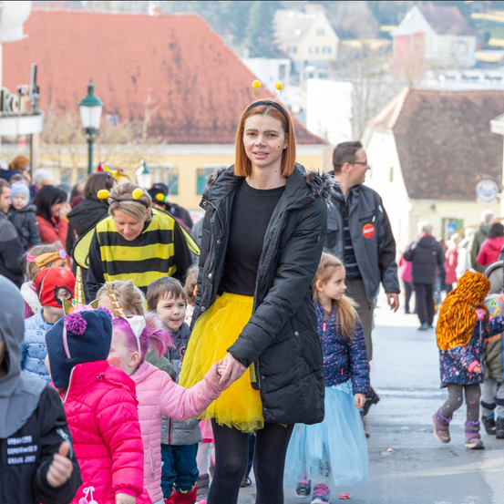 Eine Frau, die als Biene verkleidet ist, führt eine Gruppe von Kindern in einem Umzug an, umgeben von anderen in festlichen Kostümen. Im Hintergrund sind Gebäude und eine Straßenlampe zu sehen.