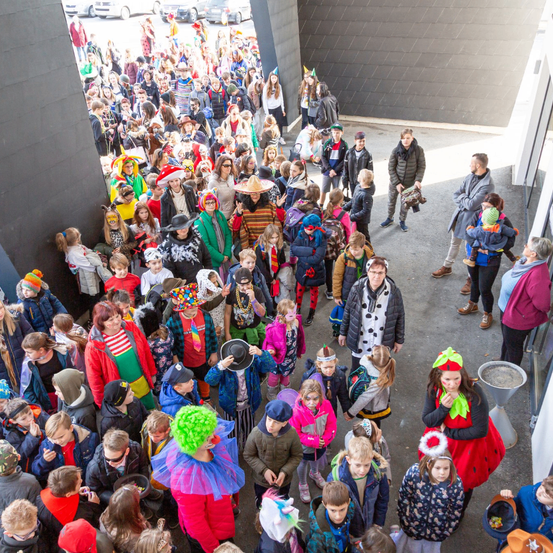 Eine große Menschenmenge, darunter Kinder in Kostümen, versammelt sich in einem Außenbereich. Einige halten Luftballons und Hüte, während andere Rucksäcke tragen. Einige Leute stehen an der Seite. Ein Mülleimer steht auf der rechten Seite.