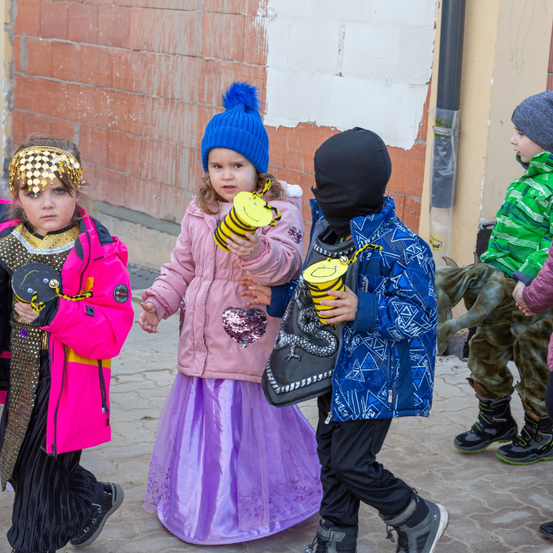 Eine Gruppe von Kindern in verschiedenen Kostümen geht auf einer Straße. Ein Kind trägt einen rosa Mantel, ein anderes eine blaue Jacke mit Gesichtsmaske.