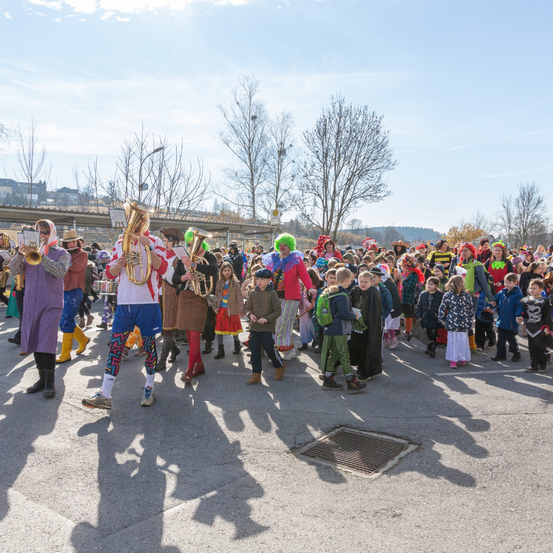 Ein fröhlicher Umzug von verkleideten Kindern und Musikern, die unter blauem Himmel marschieren, während Zuschauer sie anfeuern.