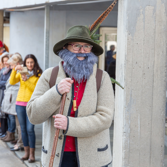 Ein Mann mit Bart und Hut steht draußen. Er hält einen Holzstab und trägt einen gestrickten Mantel. Eine Gruppe von Menschen steht hinter ihm, einige machen Fotos. Die Szene ist urban.