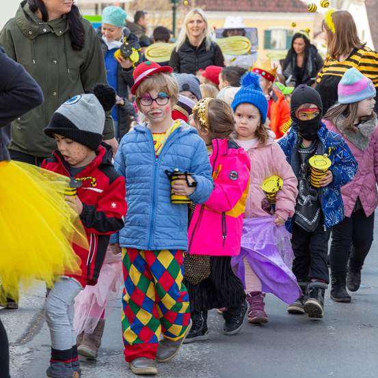 Eine Gruppe von Kindern in verschiedenen Kostümen geht bei einer Parade. Sie tragen Hüte, Brillen und halten Requisiten. Dahinter beobachten Erwachsene, einige in Kostümen, die Kinder. Die Kinder scheinen aufgeregt zu sein, und die Atmosphäre ist festlich.