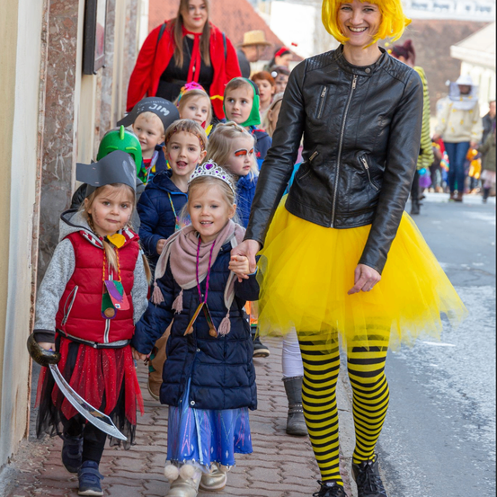Eine Frau in einem gelben Tutu und einer schwarzen Lederjacke führt eine Reihe von Kindern in Kostümen die Straße entlang. Einige Kinder tragen Piratenhüte und -umhänge, während andere Schminkfarben im Gesicht haben.