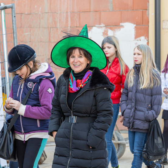 Eine Frau in einem grünen Hexenhut steht vor einer Ziegelmauer mit drei jungen Mädchen. Eines der Mädchen trägt eine lila Jacke und einen Helm, ein anderes eine rote Jacke und das dritte eine graue Jacke. Sie tragen alle Winterjacken.