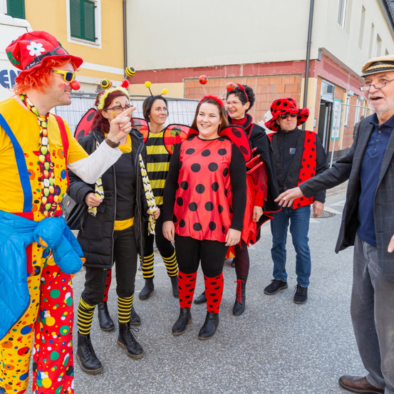 Eine Gruppe von Menschen in Marienkäfer- und Bienenkostümen steht auf einer Straße, einer zeigt mit dem Finger und die anderen lächeln. Hinter ihnen steht ein Gebäude mit einem Zaun.
