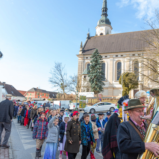 Eine Gruppe von Kindern und Erwachsenen zieht in einem Umzug vor einer Kirche. Sie tragen Kostüme und Hüte. Ein Mann spielt Tuba.