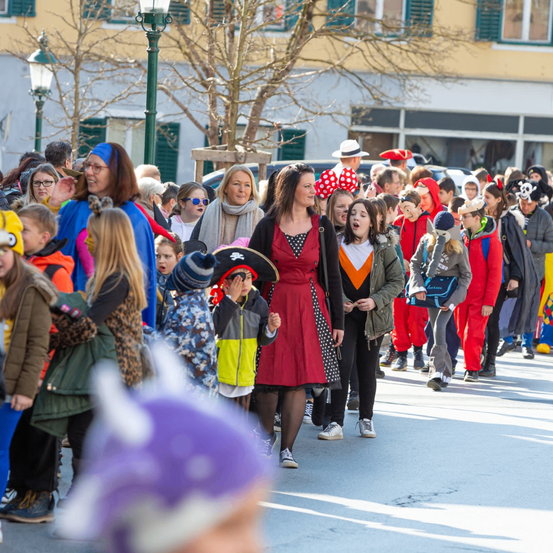 Eine Menschenmenge in Kostümen, darunter Erwachsene und Kinder, geht in einem Umzug die Straße entlang. Gebäude und Bäume säumen die Straße.