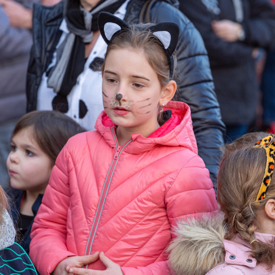 Ein Mädchen mit Katzenohren und Katzengesichtsmalfarbe trägt eine rosa Jacke und steht inmitten anderer Kinder bei einer öffentlichen Versammlung.